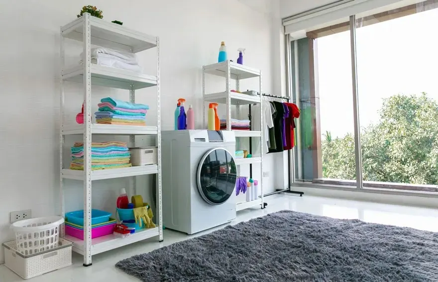 Une buanderie lumineuse avec une machine à laver, des serviettes colorées sur des étagères, des produits de nettoyage et une grande fenêtre. Marque : aucune visible.