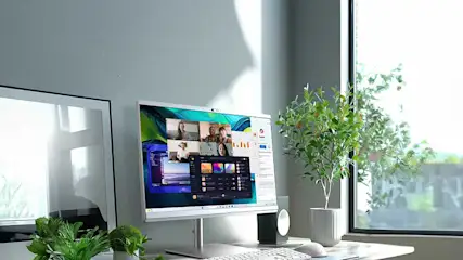 Bureau gris clair avec ordinateur, clavier blanc, souris, plante en pot et écran affichant une visioconférence.