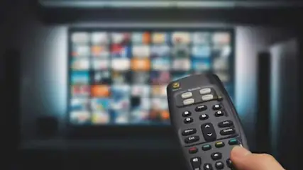 Télécommande noire au premier plan, écran TV diffusant du contenu varié en arrière-plan.