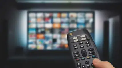 Zwarte afstandsbediening in een hand, gericht op een tv-scherm met veelkleurige miniaturen.