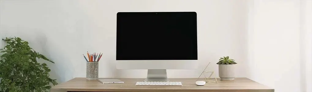 Apple iMac sits centrally on a wooden desk with keyboard, mouse, plants and pen holder; Apple logo visible beneath the screen