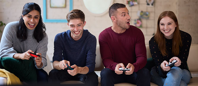 Quatre personnes assises sur un canapé jouent à des jeux vidéo avec des manettes. L'arrière-plan est un mur beige.