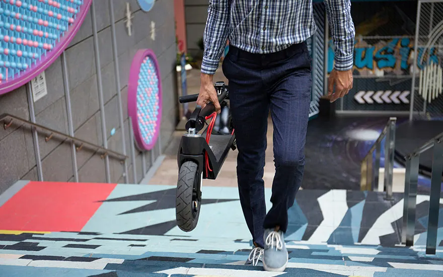 Un homme transporte une trottinette pliée dans un escalier décoré. Les murs ont des motifs géométriques et circulaires colorés.