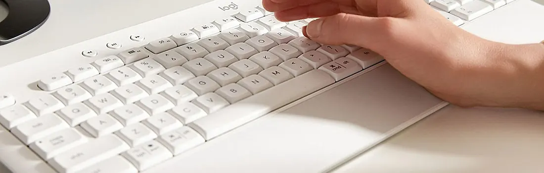Een witte Logitech toetsenbord op een bureau met een polssteun. Een hand gebruikt het toetsenbord.