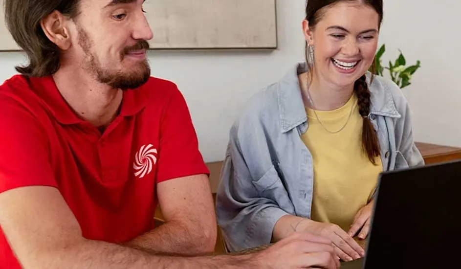 Two people at a desk look at a black laptop. One wears a red shirt with a white swirl logo, the other a yellow shirt and gray jacket.