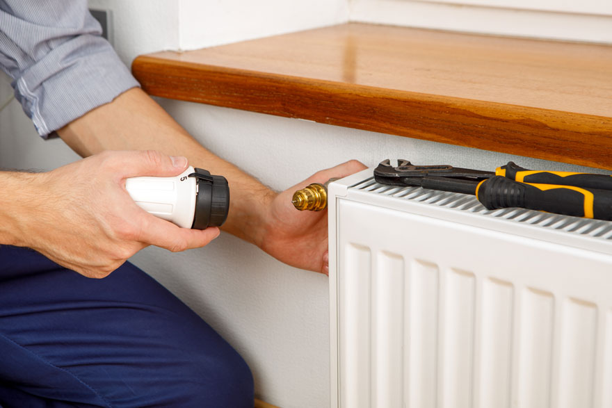Un homme ajuste un radiateur blanc avec une vanne dorée. Une clé noire et jaune repose dessus, près d'une étagère en bois.