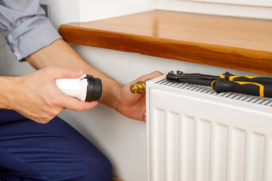 Un homme ajuste un radiateur blanc avec une vanne dorée. Une clé noire et jaune repose dessus, près d'une étagère en bois.