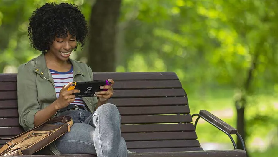 Une console de jeu portable avec des manettes colorées est tenue sur un banc en bois dans un parc verdoyant.