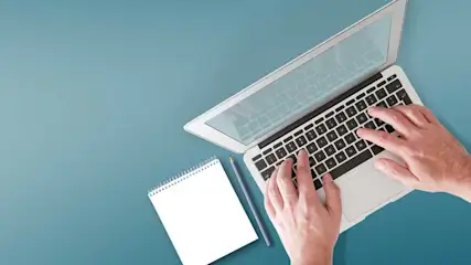 Een laptop op een blauwe tafel met een notitieblok en potlood. De handen typen op het toetsenbord.