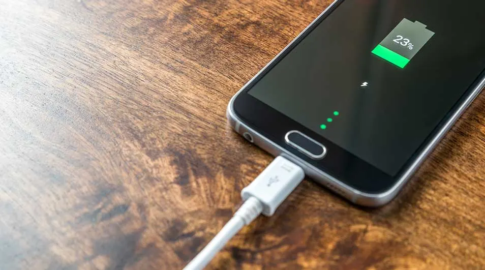 Un smartphone noir est en charge sur une table en bois, affichant une batterie à 23%.