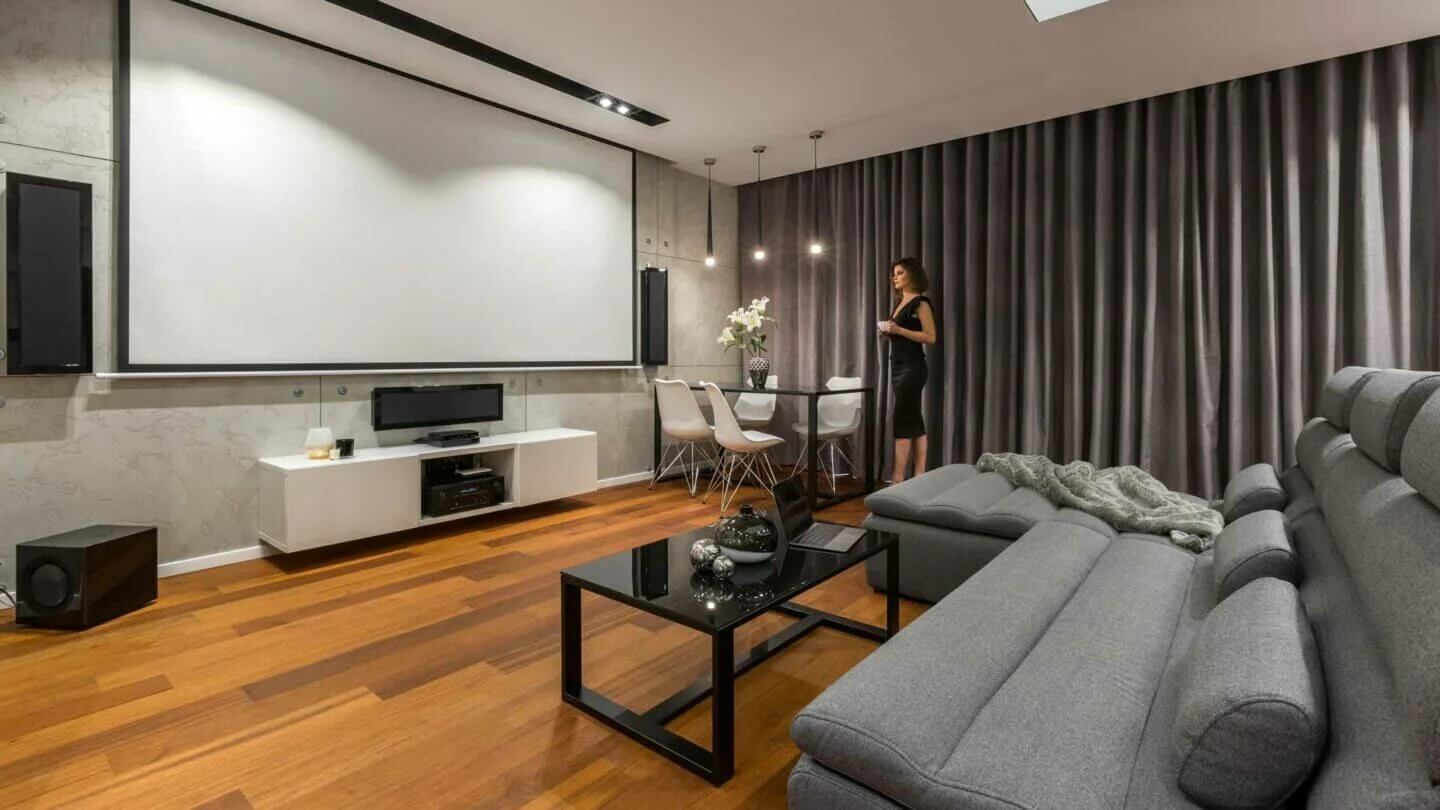 Woonkamer met grijze gordijnen, houten vloer, grijze zetel en wit tv-meubel onder een groot projectiescherm. Een zwarte tafel staat centraal.