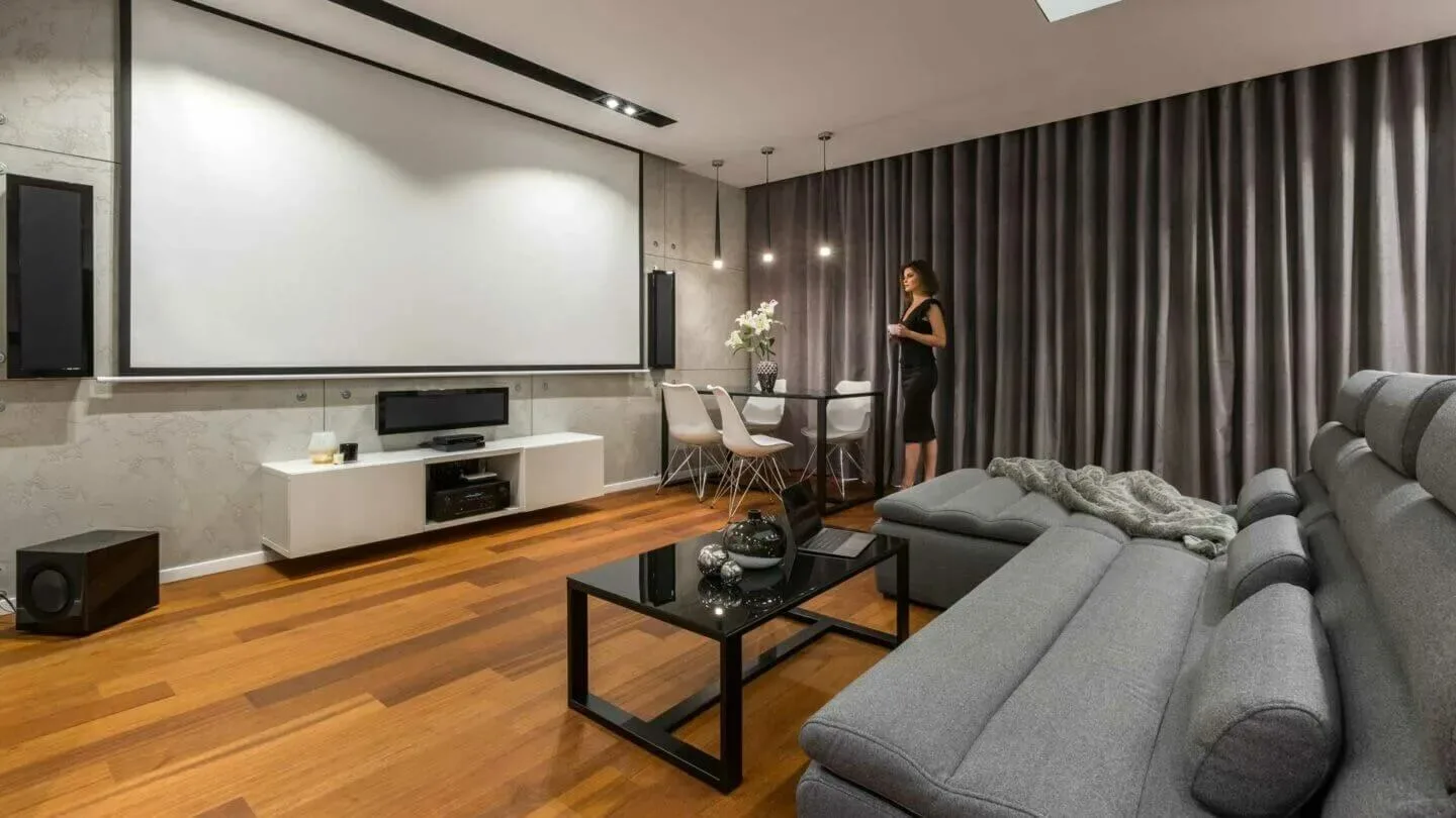 Woonkamer met grijze gordijnen, houten vloer, grijze zetel en wit tv-meubel onder een groot projectiescherm. Een zwarte tafel staat centraal.