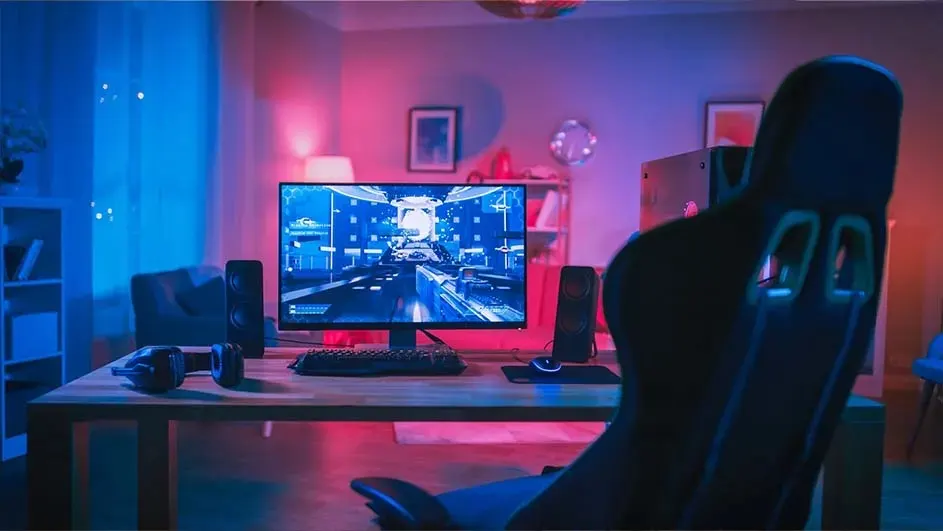 Een gamingopstelling met een bureau waarop een monitor, toetsenbord, muis en luidsprekers staan, verlicht door blauw en roze licht.