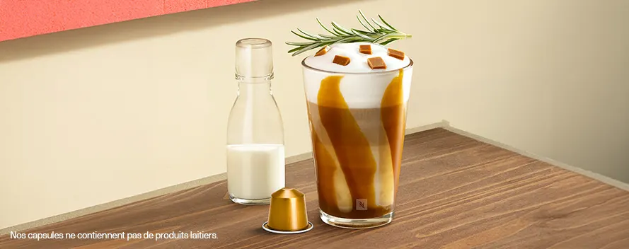 Un café glacé crémeux avec sauce caramel, romarin et morceaux de caramel dans un grand verre, à côté d’une bouteille de lait et d’une capsule dorée sur une table en bois. Une note en bas indique que les capsules ne contiennent pas d’ingrédients laitiers.