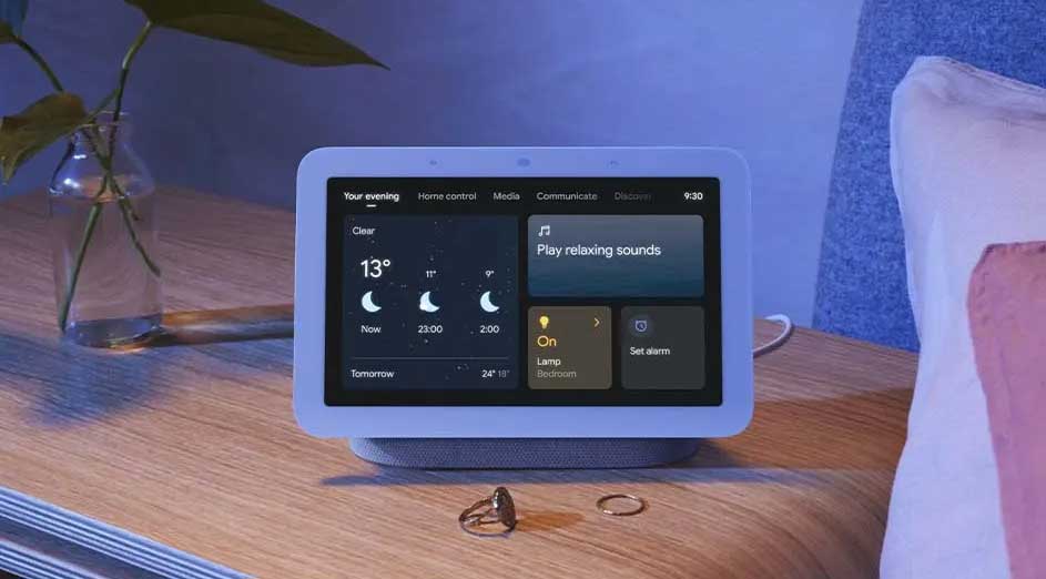 Een Google Nest Hub staat op een houten nachtkastje, met een vaas met planten en ringen. Het scherm toont het weer en slimme bedieningselementen.