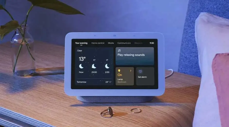 Een Google Nest Hub staat op een houten nachtkastje, met een vaas met planten en ringen. Het scherm toont het weer en slimme bedieningselementen.