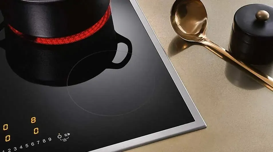 Vue rapprochée d'une plaque de cuisson noire avec une casserole, une louche dorée et un récipient noir. La plaque est allumée.