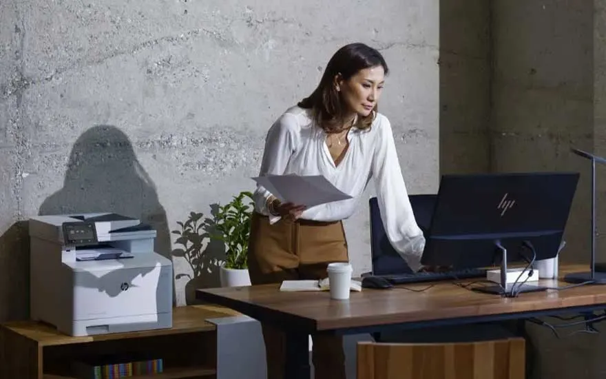 Een vrouw staat aan haar bureau met documenten in de hand en gebruikt een computerscherm. Naast haar staat een printer met papier.