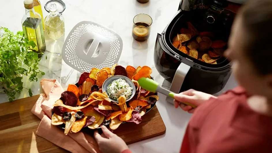 A spread of veggie chips with dip next to an air fryer, bottles and herbs on a marble surface.