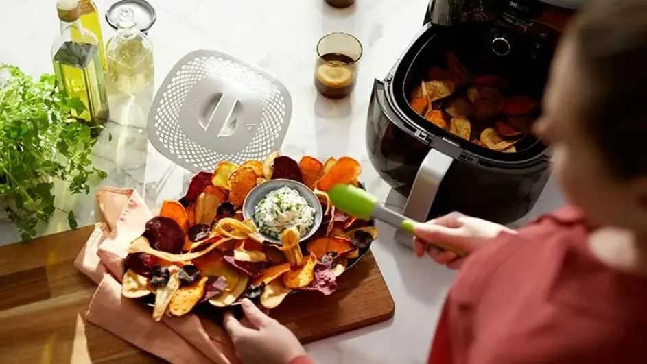 A spread of veggie chips with dip next to an air fryer, bottles and herbs on a marble surface.