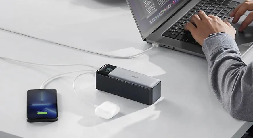 Een bureau met een laptop in gebruik, een powerbank, oortjes en een smartphone die aan het opladen is.