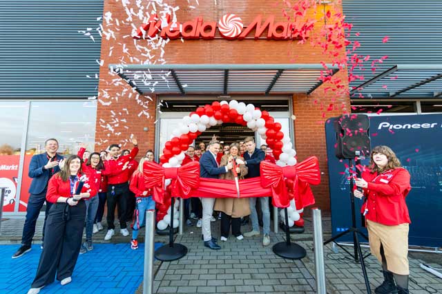 Team in rode jassen knipt rode lint door bij MediaMarkt-opening, onder boog van ballonnen met confetti in de lucht.