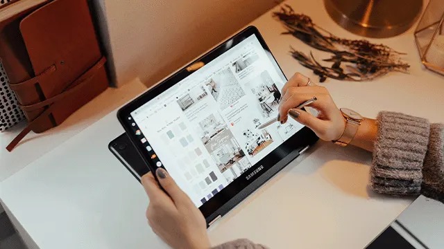 Une tablette noire affiche une page web. Des mains tiennent un stylet sur l'écran. Un agenda brun est visible à gauche.