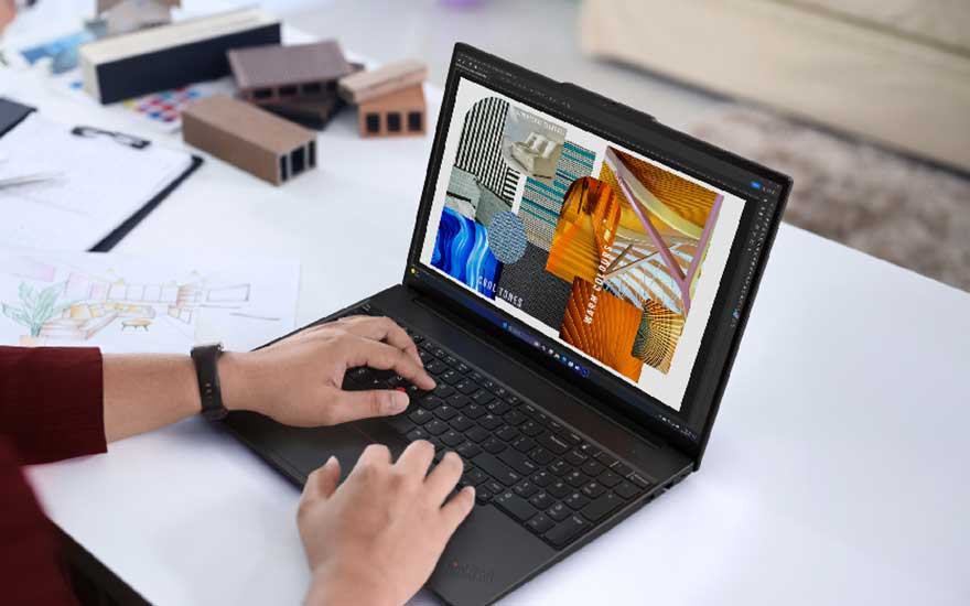 Une personne travaille sur un ordinateur Lenovo affichant des créations colorées, entourée de maquettes, dessins et fournitures de bureau.