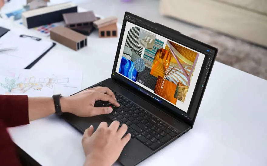 Une personne travaille sur un ordinateur Lenovo affichant des créations colorées, entourée de maquettes, dessins et fournitures de bureau.