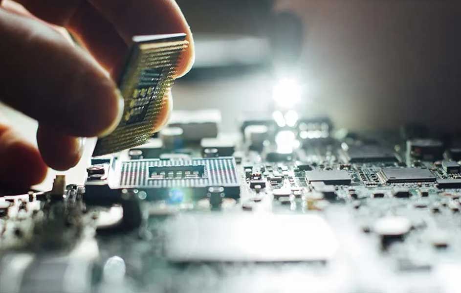 Een hand houdt een zilverkleurige computerchip boven een circuitbord vol kleine elektronische componenten. Heldere verlichting accentueert het bord.