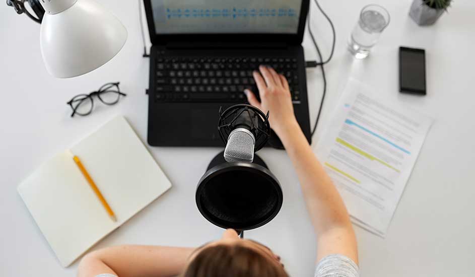 Vue de dessus d'un bureau blanc avec un ordinateur portable, un micro, des lunettes, un bloc-notes, un crayon, un verre d'eau et un smartphone.