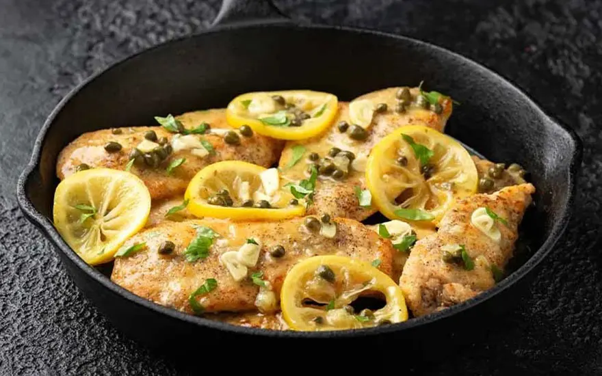 Geroosterde kip piccata in een zwarte pan met citroen, kappertjes en peterselie. Donkere achtergrond.