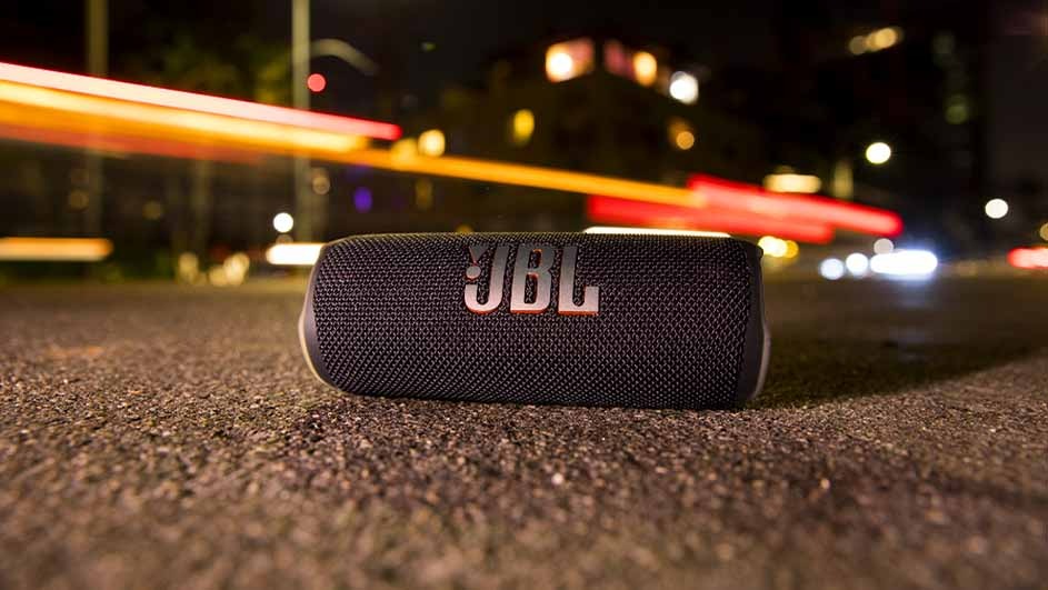 Haut-parleur JBL noir sur trottoir sombre, avec lumières et bâtiments flous en arrière-plan.