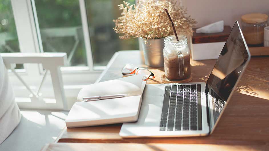 A desk with a laptop, notebook, pen, glasses, and iced coffee by a window. Dried flowers are in a metal pot.