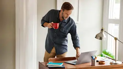 Un homme tient une tasse rouge devant un ordinateur portable sur un bureau en bois. Une lampe argentée est visible.