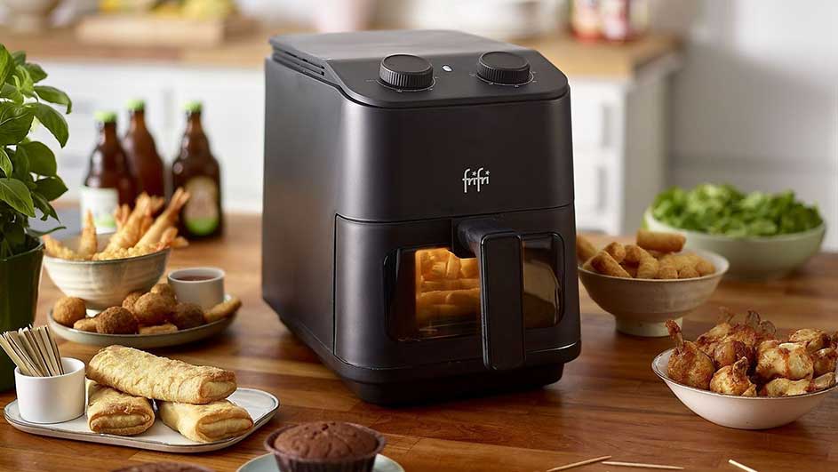 Een zwarte airfryer staat op een houten tafel, omringd door gerechten. Achtergrond: flessen en keukengerei.