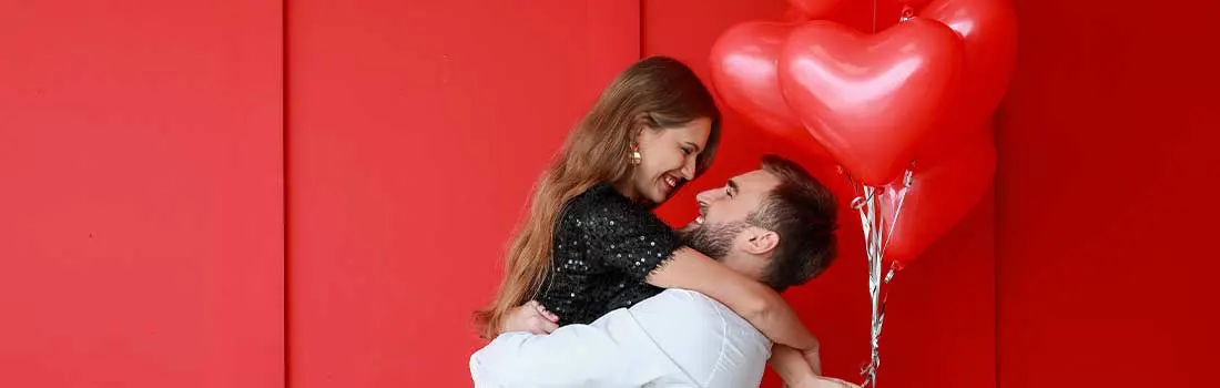 Un couple amoureux devant un mur rouge vif : il la soulève dans ses bras, ils se regardent en souriant près d’un bouquet de ballons en cœur.