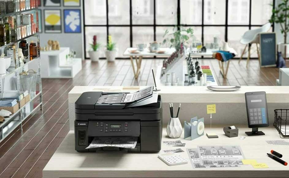 Une imprimante noire sur un bureau blanc avec des stylos, une calculatrice et une tablette. En arrière-plan, une pièce avec des fenêtres.