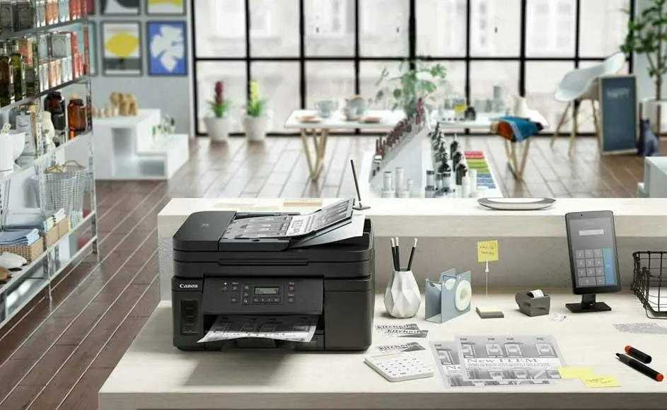 Une imprimante noire sur un bureau blanc avec des stylos, une calculatrice et une tablette. En arrière-plan, une pièce avec des fenêtres.