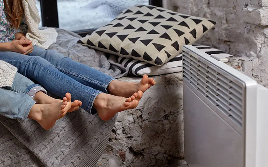 Twee paar voeten in jeans liggen op een grijze deken bij een witte radiator. Kussens met patronen liggen op de achtergrond.