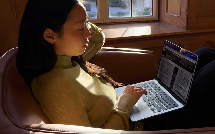 Een vrouw in een gele trui ligt ontspannen in een leren fauteuil bij het raam en leest geconcentreerd informatie op haar laptop.