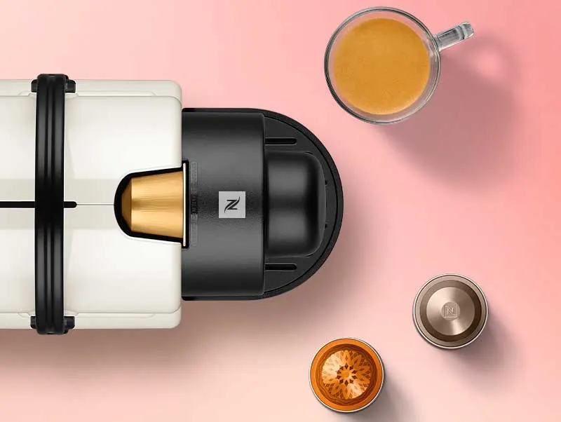 Een koffiemachine met een gouden capsule, een kop koffie en twee capsules op een roze achtergrond.