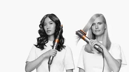 Twee personen gebruiken een Dyson Airwrap op een witte achtergrond. Het apparaat is zilver met een koperen detail.