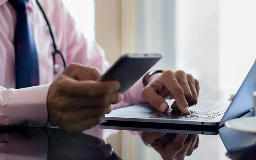 Een persoon in een roze overhemd met een stethoscoop gebruikt een laptop en houdt een zwarte smartphone vast.