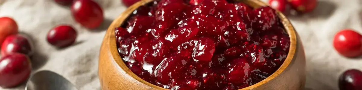 Un bol en bois rempli de sauce aux canneberges rouge vif est posé sur un tissu texturé, avec des canneberges fraîches éparpillées.