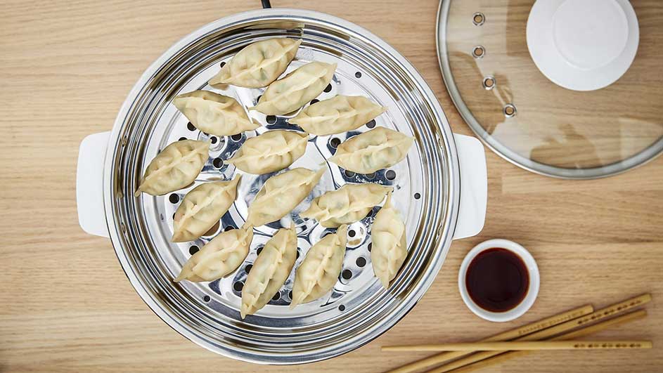 Gestoomde dumplings in een metalen stoomkoker, met sojasaus en stokjes op een houten tafel.