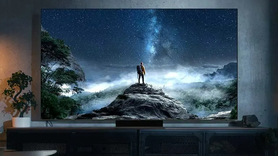 Une télévision Samsung affiche deux personnes sur un sommet de montagne sous un ciel étoilé, entourées d’arbres brumeux et de montagnes dans une pièce sombre.