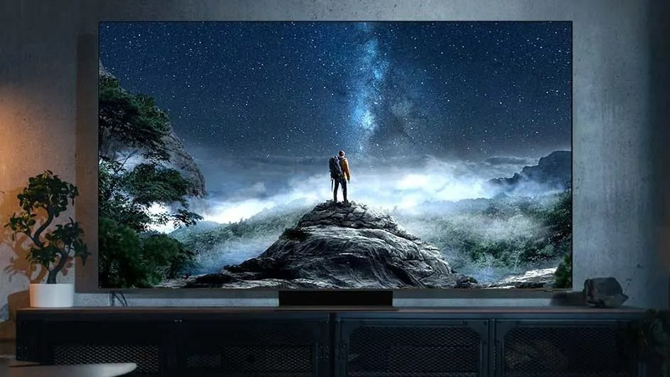 A Samsung TV displays two people on a mountain peak under a bright starry sky, surrounded by misty trees and mountains in a dark room.