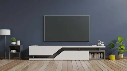 Een woonkamer met een grijze tv aan een blauwe muur, een witte kast, een grijze bijzettafel, een witte lamp en houten vloeren.