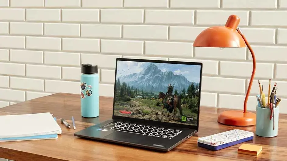 Een bureau met een laptop met een spel erop, een oranje lamp, een fles, een notitieboek en pennen.